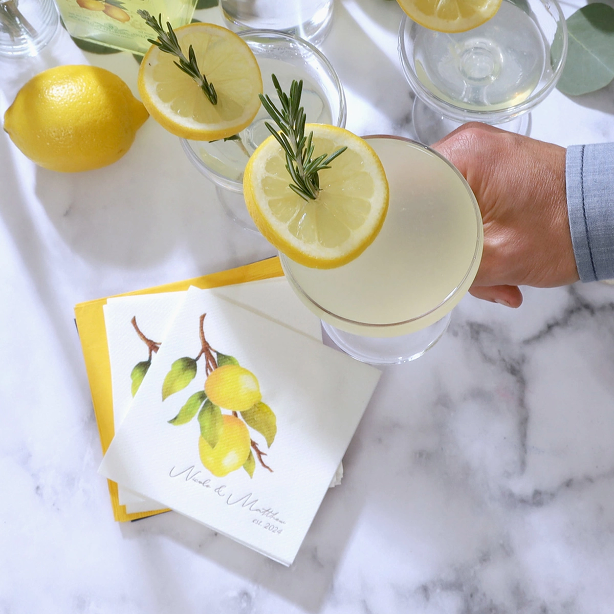 Hand holding a limoncello martini. A custom cocktail napkin with a lemon on it in the background
