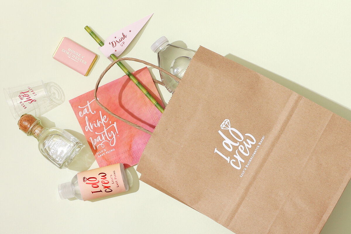 Brown goodie bag that says “I Do Crew” on it with products spilling out of it such as water, a small bottle of alcohol, a shot glass, straw, etc.