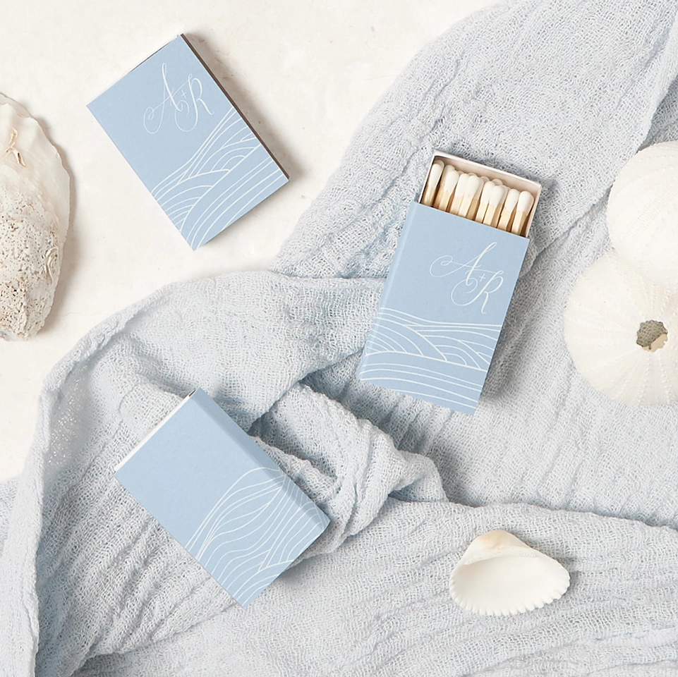 Three light blue matchboxes with wave pattern, surrounded by linen fabric and sea shells 