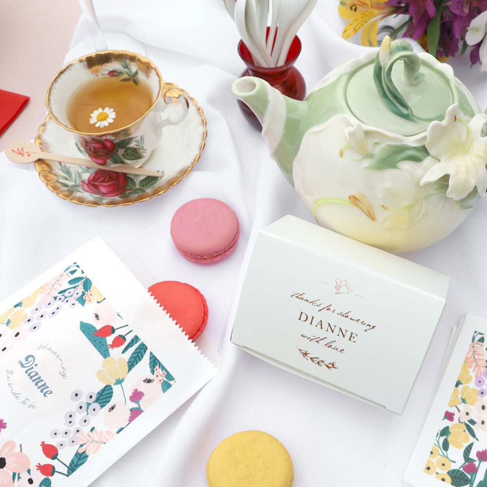 Table with tea cup and teapot along with a cake box and cookie bag with macarons surrounding it