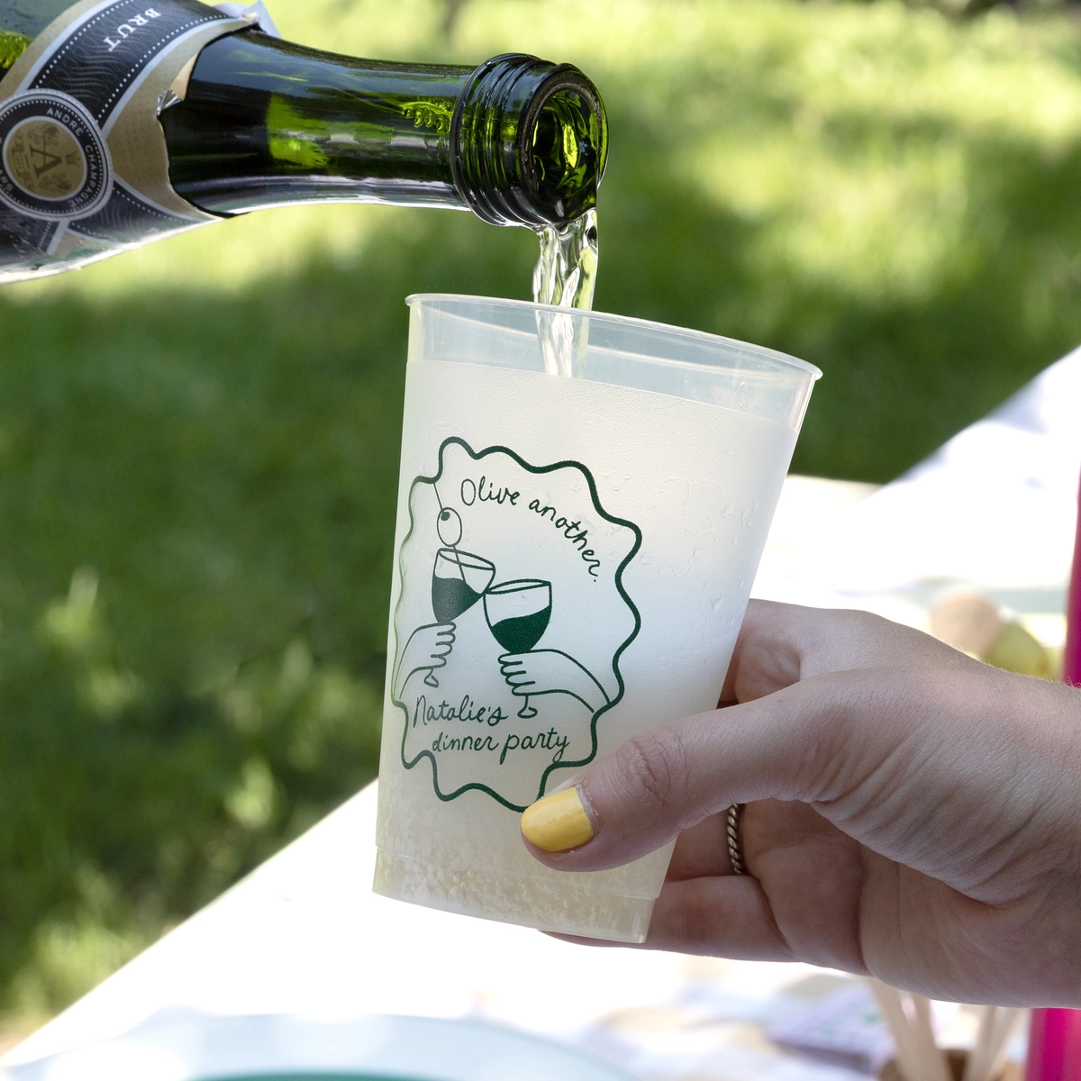 Champagne being poured into a frosted cup that says "Olive another" with a graphic of two wine glasses clinking