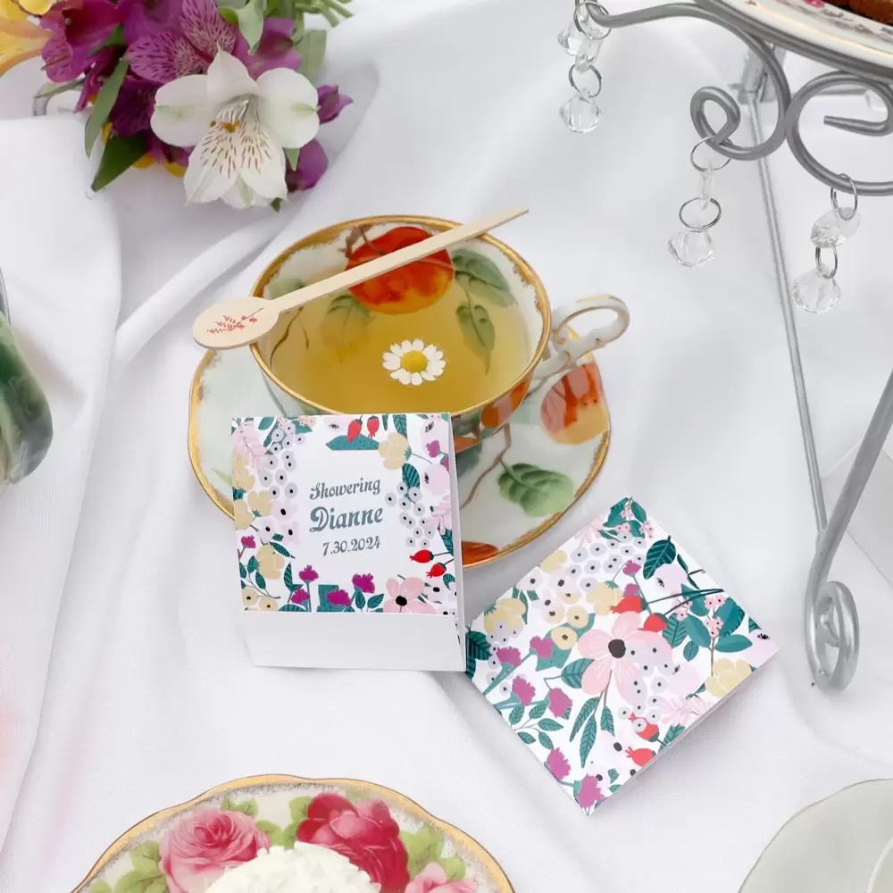 Teacup filled with tea with a stir stick laying across the tea cup. Two tea favors sitting next to the cup  