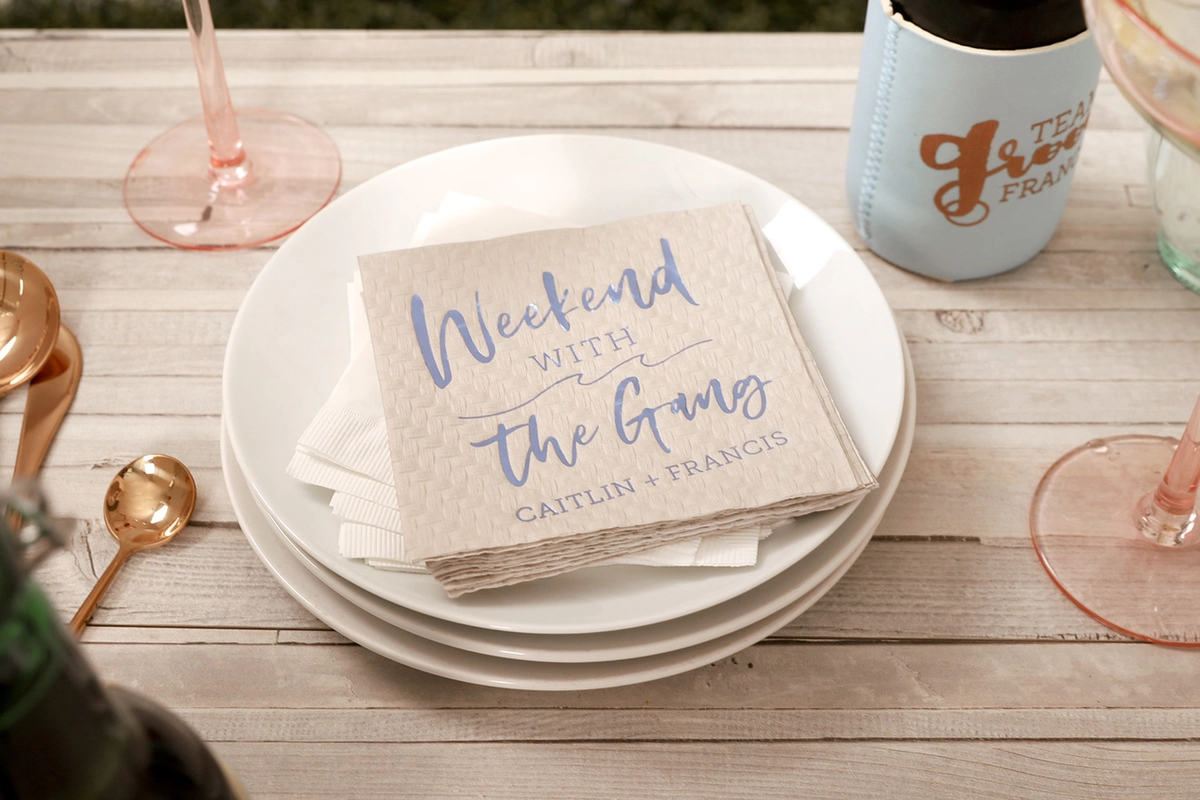 Cream textured napkin on top of a plate that says “Weekend with the Gang” in blue writing. 