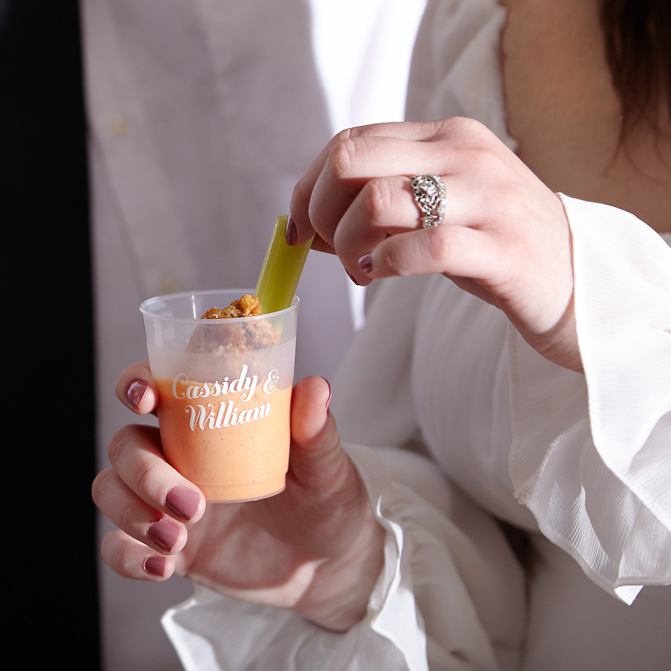 Custom plastic shot glass with 2 names printed on it. The shot glass is filled with sauce and is shown with a hnd dipping a chicken wing into the cup.