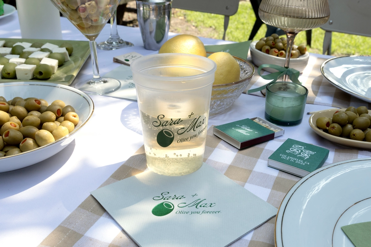 Outdoor table with patterned table cloth and a cup and napkin that have an olive graphic on them that both say "Sam + Max Olive You Forever" 