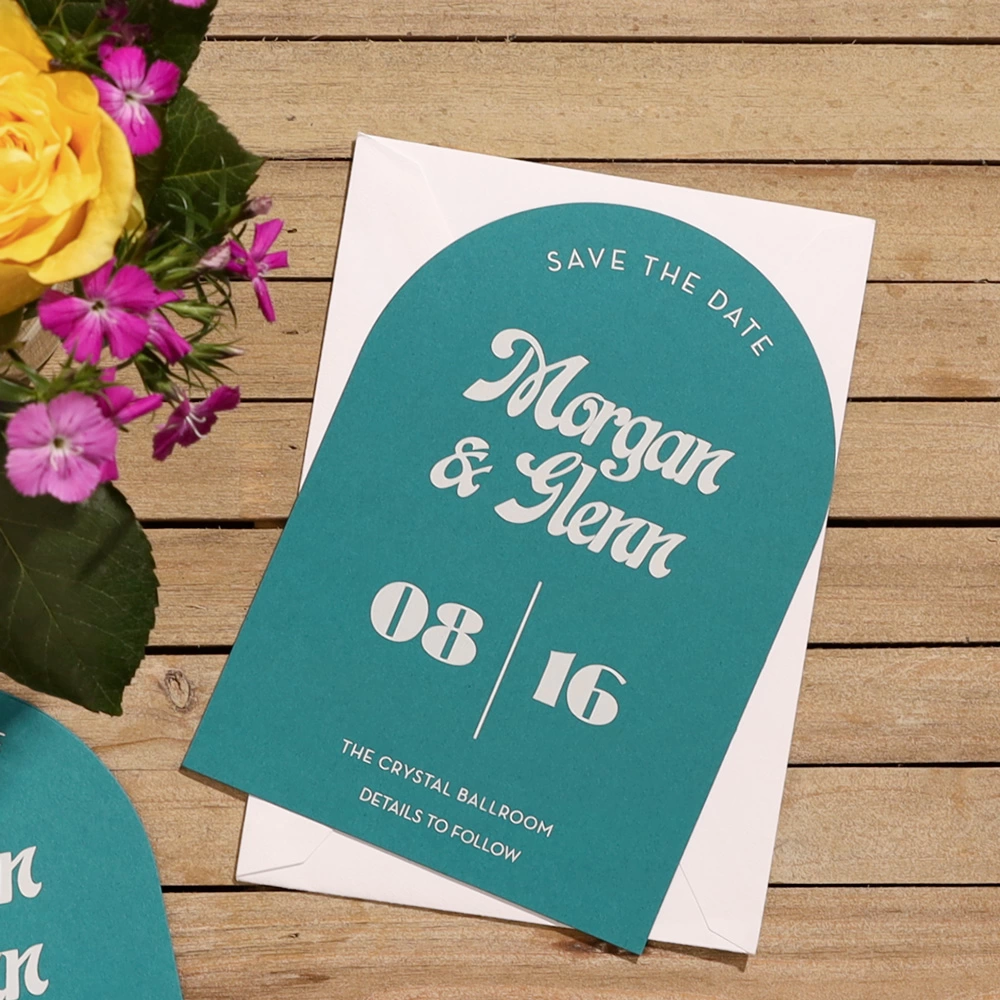 A teal minimal arch invitation on a picnic table with a pen