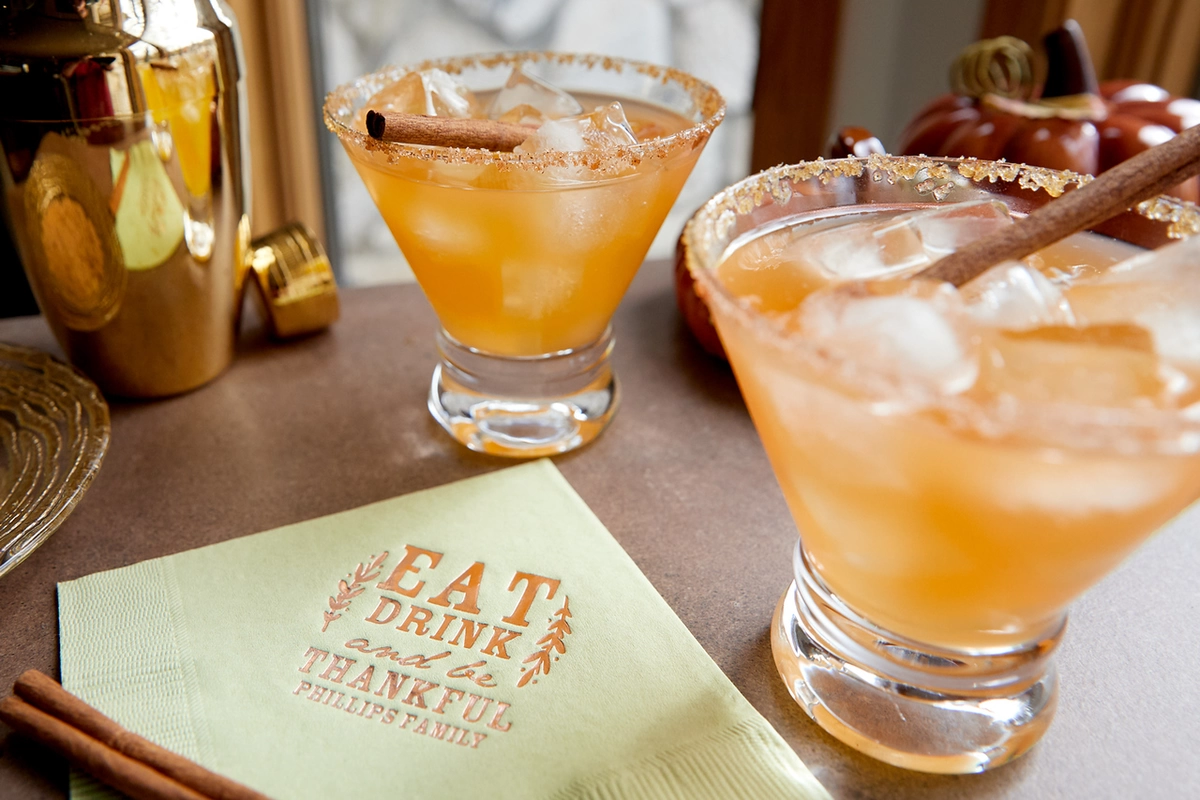Cocktails that look like cider in glasses next to a light green napkin that says "Eat, Drink, and be Grateful" 